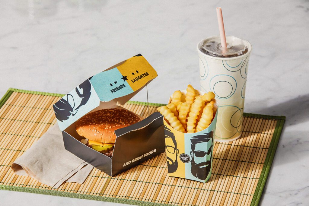 Abbildung mit einem Burger in einer faserbasierten Doppelschale, einer Pommes-Verpackung und Kaltgetränkebechern in einem Schnellrestaurant.